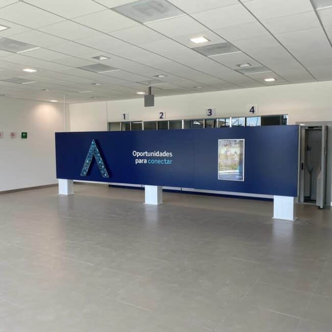 Zona de cajeros automáticos en una sucursal de BBVA con pared curva azul corporativo, tres terminales integradas y señalética podotáctil amarilla en el suelo.