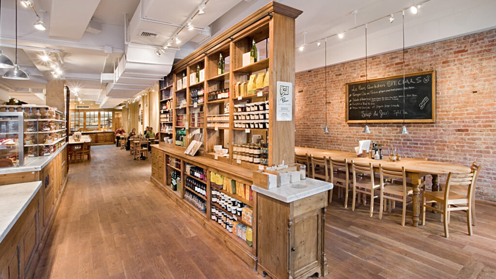 Interior de un restaurante de estilo rústico moderno con acabados en madera, una estantería central llena de productos gourmet, mesas largas de madera clara y una pared de ladrillo visto con una pizarra de menú.